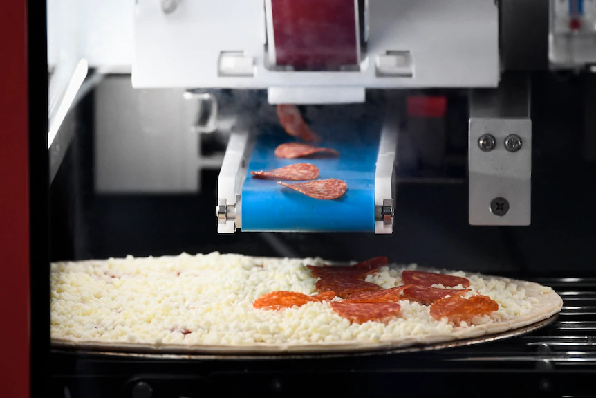 A conveyer belt drops salami on a cheesy uncooked crust